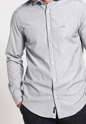 Homme portant une chemise à manches longues boutonnée gris clair avec le logo "AJ" sur la poitrine, les mains dans les poches, et un pantalon noir.