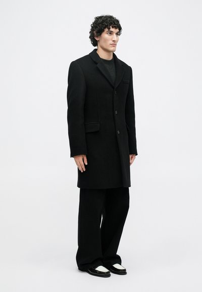 Άνδρας με σγουρά μαλλιά ντυμένος με μαύρο μακρύ παλτό, μαύρο φαρδύ παντελόνι και μαύρα-άσπρα λουστρίνια, στέκεται μπροστά σε απλή λευκή επιφάνεια.