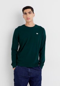 Langsleeve, donkergroene shirt gemaakt van soepel stof. Heeft een klein logo op de borst, ronde halslijn en smalle manchetten. Gecombineerd met denim.