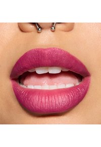 Rouge à lèvres fuchsia mat appliqué sur des lèvres pleines, présentant une texture lisse et une forme définie. Détails dentaires visibles et un piercing à la narine.
