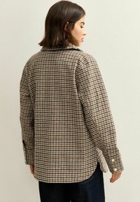Brun rutig flanellskjorta med mjuk textur, knäppning vid ärmsluten och krage. Designen har ett rutmönster i dämpade toner.