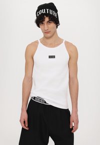 Hvit ribbet tank topp med en svart etikett som leser "COUTURE" og svarte løstsittende bukser med Versace-logo på midjebåndet. Svart beanie med hvit tekst.