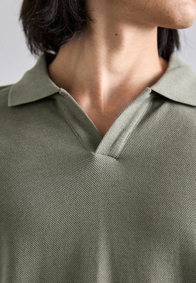 Polo vert olive avec un tissu texturé, un col classique et un détail de cou en V unique. Design décontracté et minimaliste.