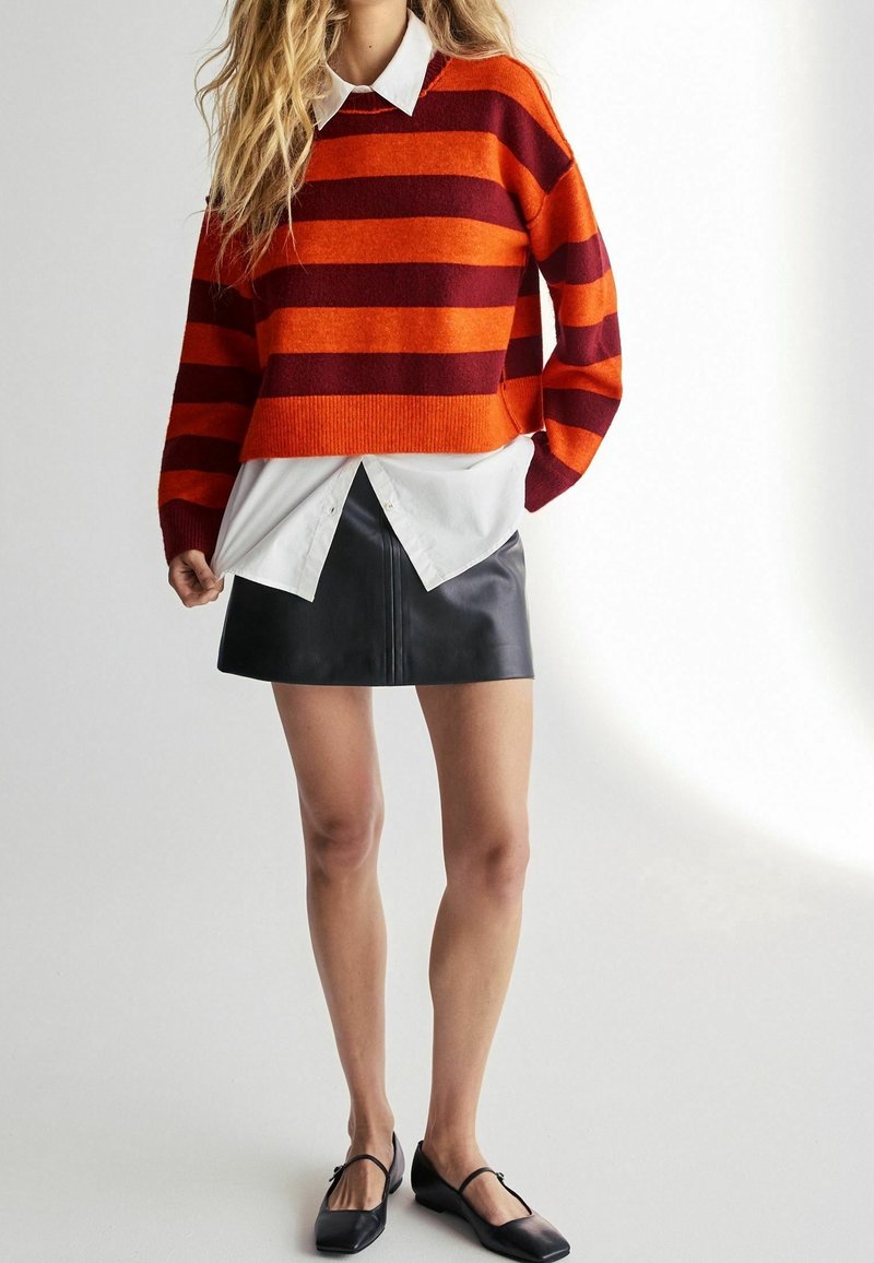 Gestreepte trui in rood en oranje, gelaagd over een witte blouse met kraag, gecombineerd met een zwarte leren minirok en zwarte ballerina's.