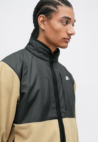 Černá a béžová zipová bunda s vysokým límcem, vyrobená z fleecu a nylonu. Obsahuje logo Nike a kapsu na zip.