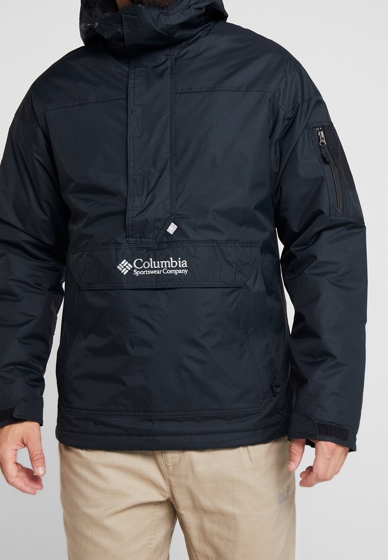 Homme portant une veste à capuche noire Columbia Sportswear avec poche avant et pantalon beige sur un fond uni.