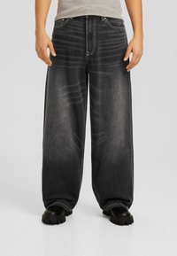 Jeans de mezclilla oscuros y holgados con una textura desgastada y corte recto. Con cinco bolsillos y cierre de cremallera. Botas gruesas de color negro mate.