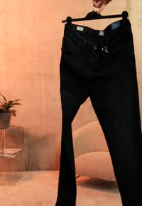 Jean noir tenu sur un cintre par une main dans une pièce aux tons chauds avec une plante sur une table en métal et un fauteuil texturé en arrière-plan.