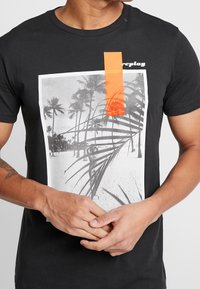 Sort bomulds t-shirt med et fotografisk tryk af palmetræer og en strand, accentueret med et orange rektangulært stykke tape.