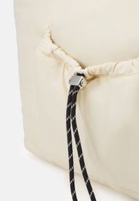 Coussin matelassé de couleur crème avec une ouverture resserrée, doté d'un cordon noir à motifs blancs et d'un ajusteur métallique.