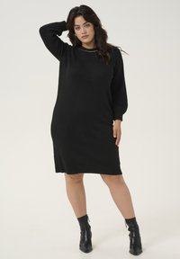 Black knit sweater dress with long sleeves, ribbed cuffs, and a round neckline. Worn with black ankle boots. Smooth texture, knee-length.