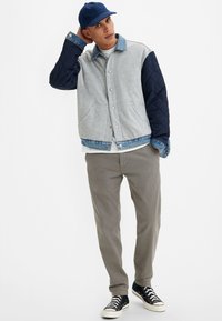 Giacca trapuntata grigia e blu navy con colletto in denim, con tasche frontali, indossata sopra una camicia bianca, abbinata a pantaloni beige e sneakers nere.