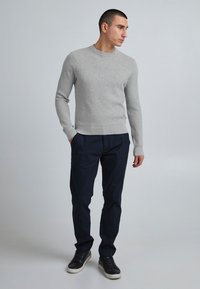 Bărbat în picioare cu mâinile în buzunare, purtând un pulover texturat de culoare gri deschis, pantaloni negri închis și adidași negri cu talpă albă.