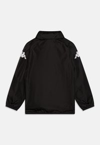 Juodas ilgam rankoviam vėjo nepraleidžiantis švarkelis su stovimu apykakliu ir baltais logotipais su dviem sėdinčiomis figūromis ant abiejų viršutinių rankovių.