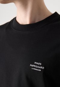 Čierne bavlnené tričko s okrúhlym výstrihom a malým bielym vyšívaným logom na hrudi. Hladká textúra, štandardný strih.