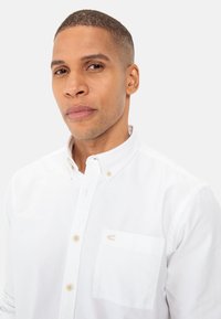 Witte button-down shirt met kraag, voorzien van een borstzak en een subtiel logo. Gemaakt van een glad materiaal, de textuur lijkt strak en schoon.