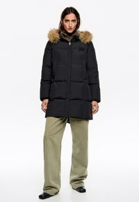 Czarna kurtka puchowa z odpinanym kapturem z beżowego sztucznego futra, zapinana na zamek, z bocznymi kieszeniami. W zestawie z oliwkowozielonymi szerokimi spodniami i czarnymi butami.