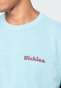 Camiseta de manga corta de color azul claro hecha de algodón. Presenta un logotipo en color marrón "Dickies" en la parte izquierda del pecho y un cuello redondo acanalado.