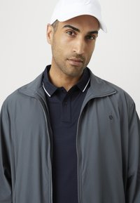 Mann som har på seg hvit caps, marineblå polo-skjorte og grå jakke med glidelås, vendt mot kameraet foran en enkel lys bakgrunn.
