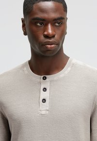 Beige gestricktes Henley-Hemd mit strukturiertem Muster, runder Ausschnitt und vier dunklen Knöpfen. Nahaufnahme des Oberkörpers.