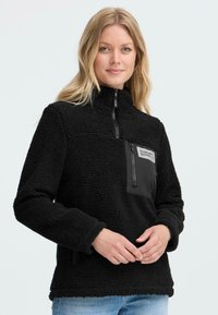 Zwarte fleece pullover met hoge kraag, halflange rits en een borstzak aan de linkerkant met een zwarte stoffen accent. Getextureerd, zacht materiaal.
