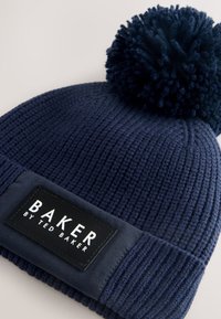 Tengerészkék kötött sapka bordázott mintával, nagy bojttal a tetején és egy fekete címkével, amelyen a "BAKER by TED BAKER" felirat olvasható.