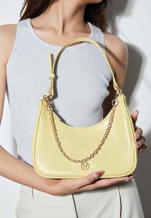 Frau trägt ein hellgraues Tanktop und hält eine blassgelbe Handtasche mit Goldketten-Detail und kleinem rundem Logo auf der Vorderseite.