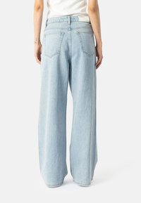 Osoba u svijetloplavim širokim trapericama s stražnjim džepovima i bijelom majicom, prikazana s leđa na bijeloj pozadini.