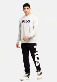 Pelēka garām piedurknēm izgatavota sporta krekls ar sarkano un zilo "FILA" logo, kombinācijā ar melniem sporta biksēm ar lielu balto "FILA" dizainu. Baltas kedas.