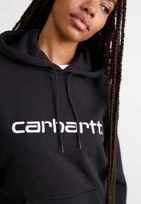 Felpa con cappuccio nera in cotone, caratterizzata da un grande logo "carhartt" bianco. Il design include una tasca frontale a marsupio e cappuccio con coulisse.