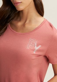 Camiseta de algodón rosa con escote redondeado y mangas cortas. Presenta un gráfico de "FY" adornado con diamantes en la parte izquierda del pecho.