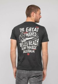T-shirt noir avec un graphique de vague et le texte : "Les Grandes Vagues Belles Beach Surf Paradis Australie" en blanc et rouge. Manches courtes.