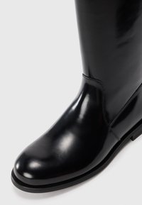Black polished leather knee-high boot with rounded toe and low heel on white background.
