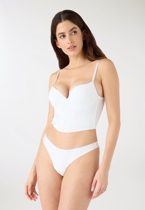 Kobieta ubrana w biały długi biustonosz i dopasowane białe majtki, stojąca na tle jednolitego jasnego tła, patrząca w bok.