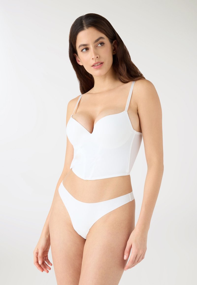 Donna che indossa un reggiseno lungo bianco e mutandine abbinate bianche, in piedi contro uno sfondo chiaro uniforme, che guarda di lato.