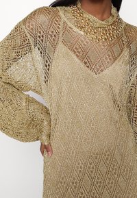 Femme portant une robe en maille métallique dorée avec des motifs complexes et un grand collier imposant orné de perles dorées et de perles.