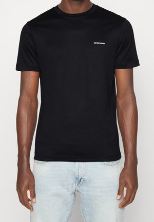 Mand iført en tætsiddende sort Emporio Armani t-shirt med lille hvidt logo på brystet, kombineret med lyseblå jeans.