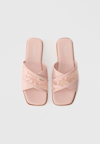 Pink slide sandals with a crisscross design, featuring a lace overlay and a smooth, matte finish. The sole is flat and minimalistic.