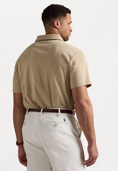 Cămașă bej cu mânecă scurtă și guler, cu un model subtil, purtată cu pantaloni deschiși la culoare ce au un mic logo albastru pe buzunarul din spate.