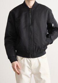 Veste bomber noire avec fermeture éclair, poignets et ourlet côtelés, poches latérales et une poche utilitaire sur la manche. Texture de tissu lisse.