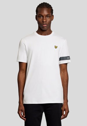 Ung mand med flettet hår iført en hvid T-shirt med en gul fuglelogo på brystet og sorte bukser mod en grå baggrund.