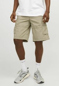 Shorts cargo beiges avec plusieurs poches latérales, coupe droite et détails de couture visibles, portés avec des chaussettes blanches et des baskets grises.