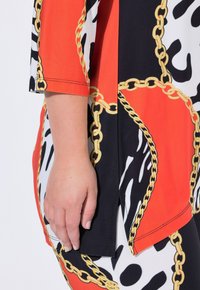Kolorowa tunika z wzorami w kolorach pomarańczowym, czarnym i białym, ozdobiona złotymi łańcuszkami. Miękka tkanina, luźny krój i rękawy do łokcia.