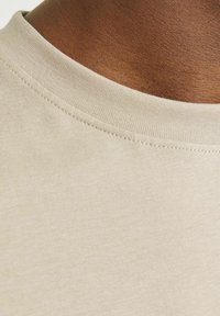 Beige Baumwoll-T-Shirt-Detail mit Rundhalsausschnitt und feiner Naht. Der Stoff hat eine glatte Textur ohne auffällige Muster.