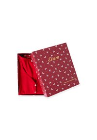 Červený oděv vystavený v vzorované krabici s bílými mašlemi. Krabice má zlatý text a květinový vzor uvnitř, který ukazuje strukturu látky.