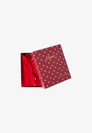 Abito rosso esposto in una scatola decorata con fiocchi bianchi. La scatola presenta un testo dorato e un design floreale all'interno, evidenziando la trama del tessuto.