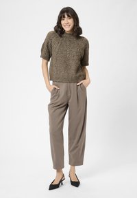 Kvinde står med hænderne i lommerne iført en brun strikket kortærmet sweater, taupe bredbenede bukser og sorte slingback hæle.