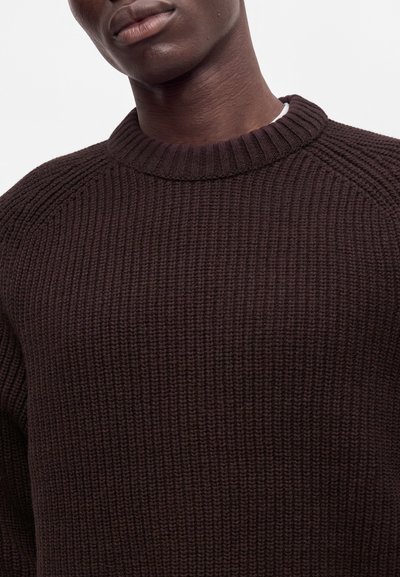 Gros plan d'une personne portant un pull en maille côtelée épaisse de couleur brun foncé avec un col rond haut sur un fond blanc uni.