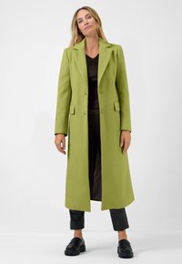 Long, open-front coat in bright green, made of textured fabric. Features notched lapels, two front pockets, and a straight fit. Black shoes below.