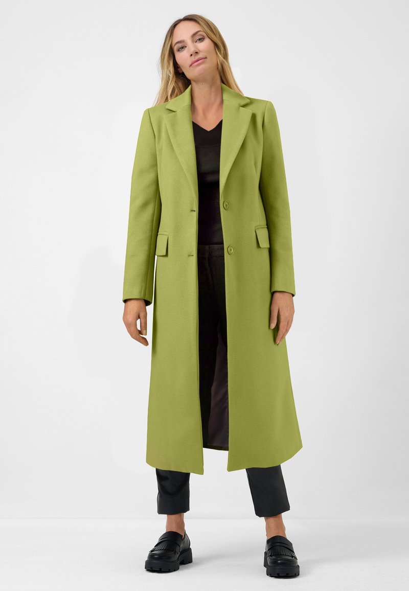 Long, open-front coat in bright green, made of textured fabric. Features notched lapels, two front pockets, and a straight fit. Black shoes below.